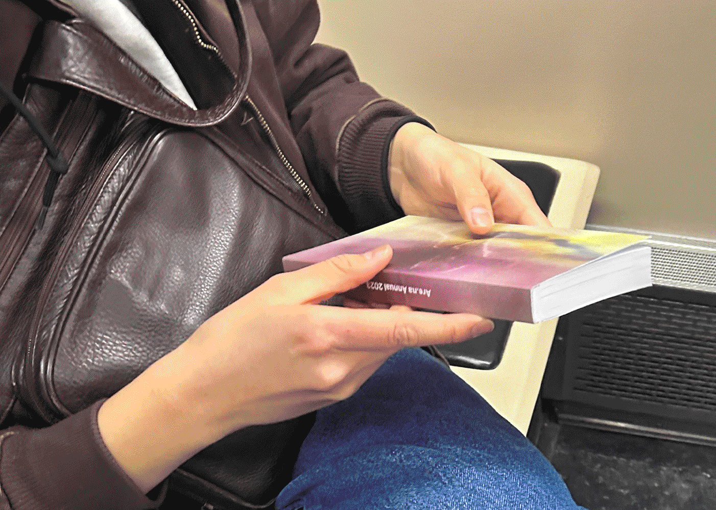 a person holding a thick, narrow book, examining the back cover. the cover is in vibrant purple and beige hues, depicting an abstract texture.