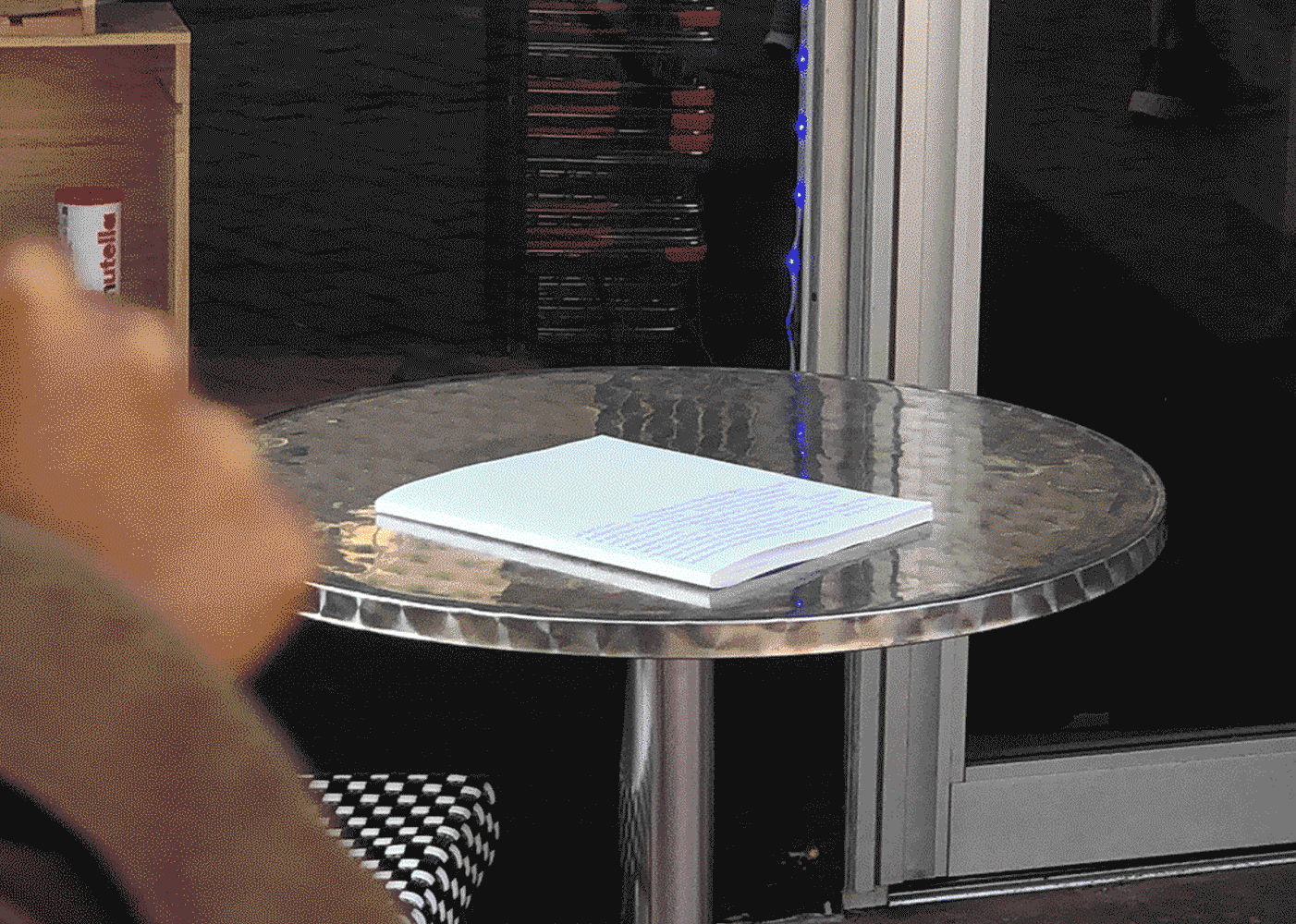 a book with a white cover on a reflective metallic tabletop in front of some shop windows. in the foreground is the hand of a stranger passing by.