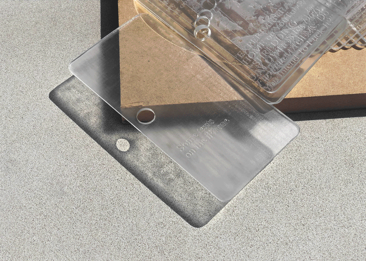sheets of clear plastic shaped like smartphones, with a hole punched in the margin to the left, displayed against a concrete background. other sheets of plastic are placed across a wooden board.